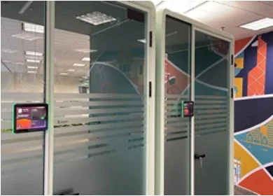 Glass meeting booths in an open-plan office, each with a booking panel at the door