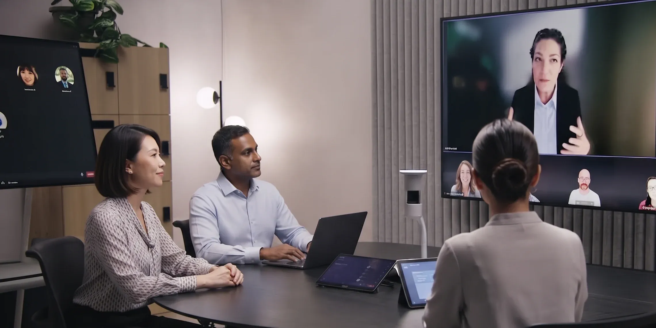 Three colleagues in a meeting room on a video call with remote participants shown on a large wall display