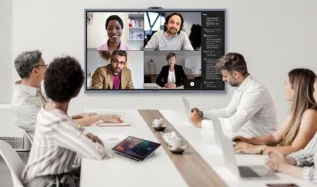 Six colleagues around a meeting room table on a video call, with four remote participants on the wall display